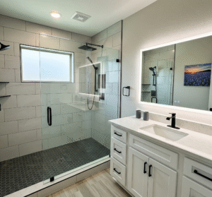 A modern bathroom remodel in a Kaufman County, Texas home, featuring a new walk-in shower with light gray subway tiles and a dark gray hexagon tile floor, a glass enclosure, a new white vanity with a quartz countertop, and a large mirror with built-in LED lighting by Affordable 1 Day Baths